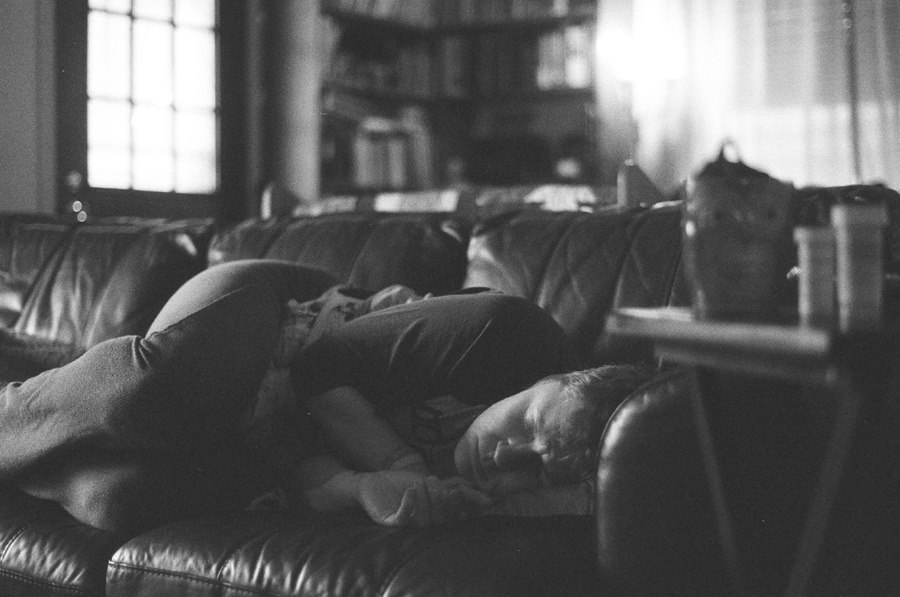 Woman lying on couch, holding heating pad to stomach, bottle prescription pain pills in the foreground next to a mug of tea