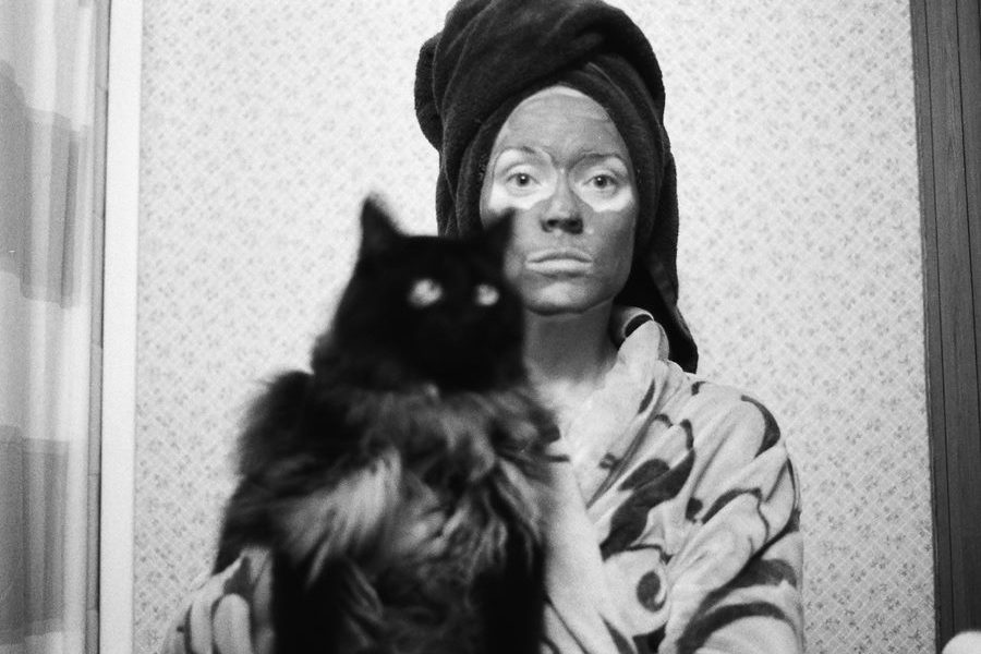 Black and white photo of a woman in bathrobe, wearing a towel on her head, clay mask on her face, holding a cat in the bathroom
